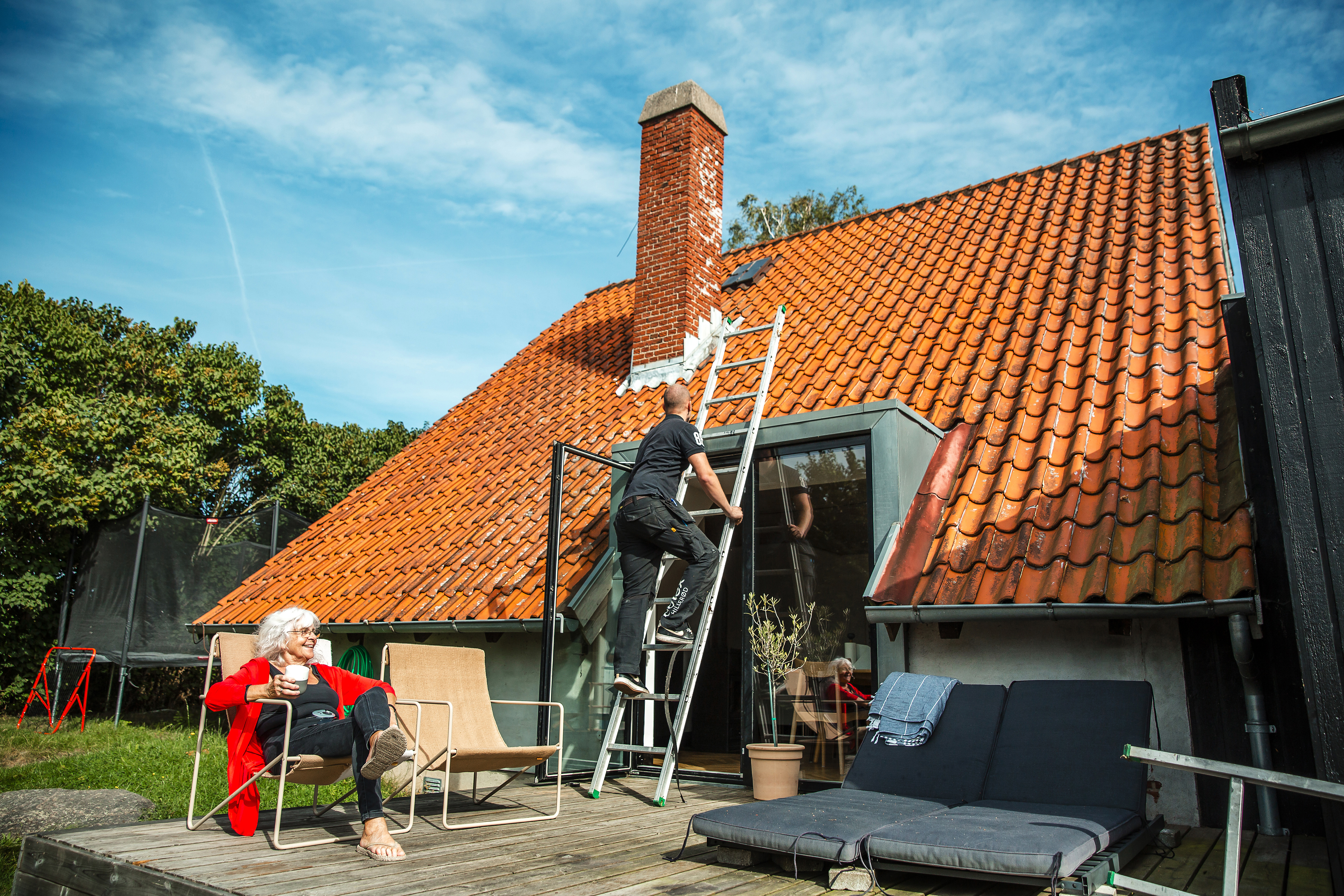 Kvinde sidder på terrasse foran hus, mens håndværker på stige arbejder på taget, fotografi