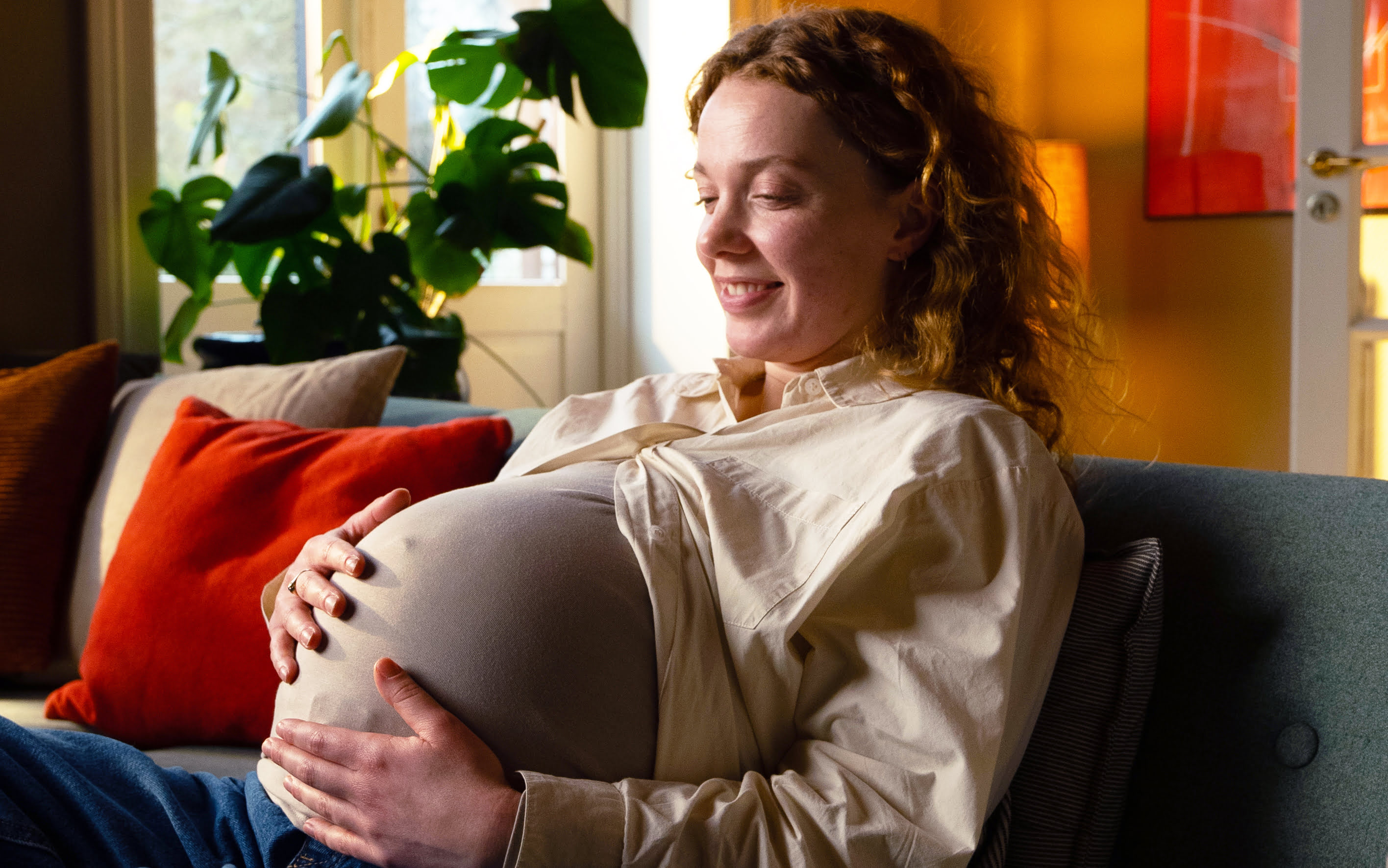 Gravid kvinde sidder i sofa, kigger på sin mave og smiler, fotografi