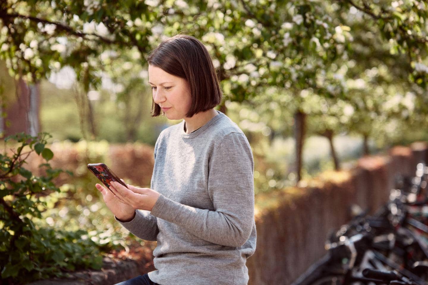 Kvinde står under træer og kigger på sin telefon, fotografi
